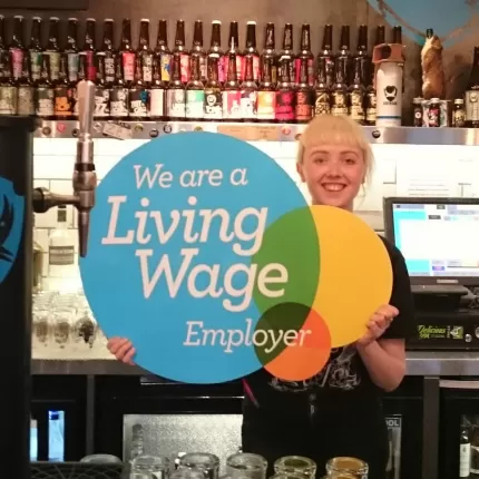 Brewdog employee posing with the Living Wage Employer logo behing the bar