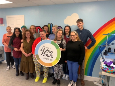 A group of Molke workers stood in front of a blue wall with a rainbow on. The workers are holding a plaque that reads 'We are a Living Hours employer'.  