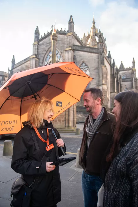 Mercat tour guide stood under an umbrella with two tourists next to St Giles Cathedral in Edinburgh, all smiling