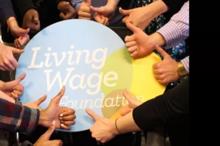 Living Wage Foundation foamboard surrounded by a circle of hands giving it the thumbs up
