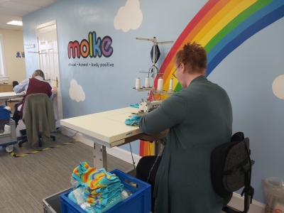 A woman sits at a white desk working on a sewing machine, in front of a blue wall with Molke's loho and a bright rainbow