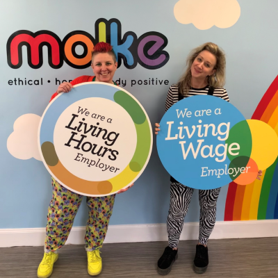 Two workers holding plaques that read 'We are a Living Hours employer' and ' We are a Living Wage employer'. The workers are stood in front of a blue wall decorated with clouds, rainbows and Molke's logo.