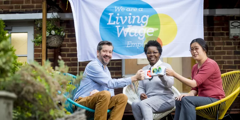 Three people sat on colourful outdoor chairs hold up Living Wage branded mugs in front of a banner with the Living Wage Foundation logo